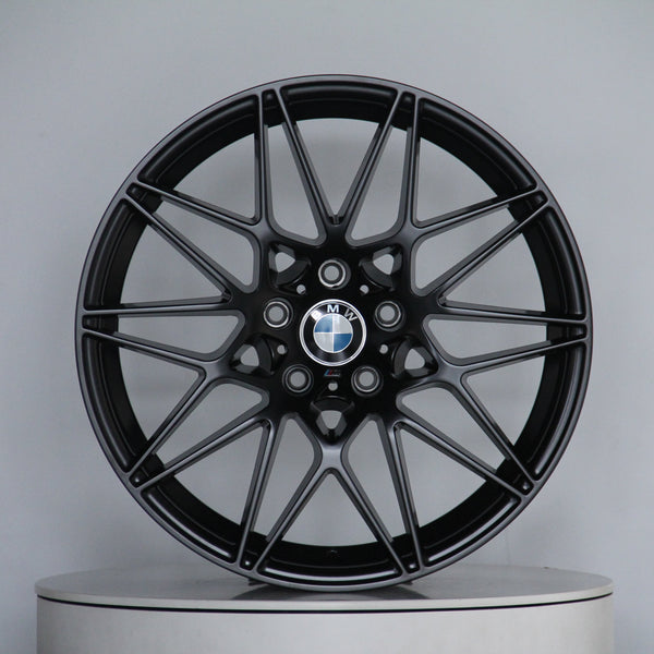 A black alloy wheel with multi-spoke design, featuring a central BMW logo, rests on a round display platform against a plain gray background.