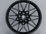 A black alloy wheel with intricate spoke design displays a BMW logo at the center, resting on a round platform against a plain gray background.