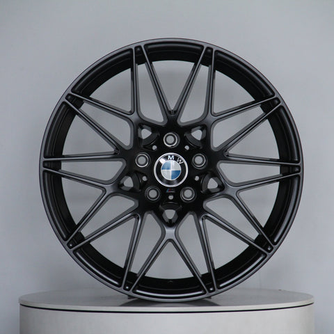 A black alloy wheel with intricate spoke design displays a BMW logo at the center, resting on a round platform against a plain gray background.