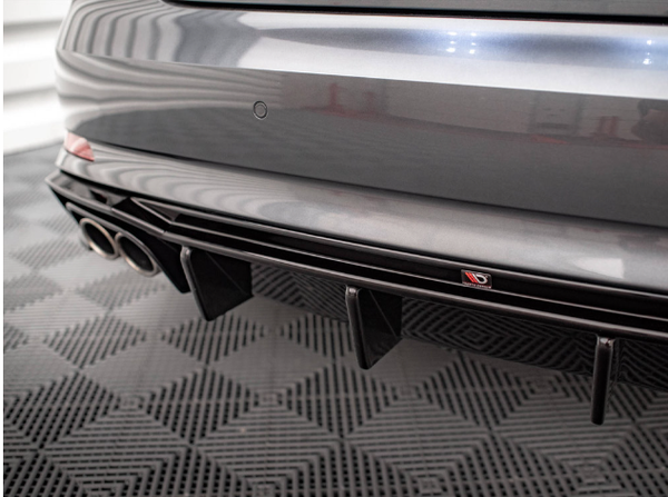 A car's rear bumper with dual exhaust pipes and a diffuser. The car is parked on a textured, black floor, and there's a small emblem displaying a red “D” and “GB”.