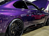 A sleek, purple sports car with a raised hood is displayed indoors on a polished concrete floor, featuring glossy black wheels.