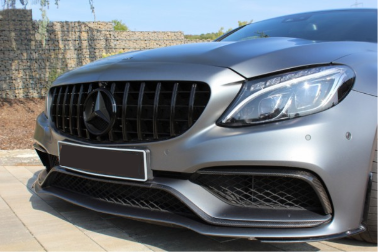 A silver car's front, displaying a prominent grille and sleek headlights, is parked on a stone driveway. The background shows a stone wall and clear blue sky.