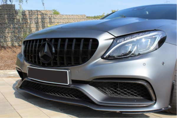A silver car's front, displaying a prominent grille and sleek headlights, is parked on a stone driveway. The background shows a stone wall and clear blue sky.