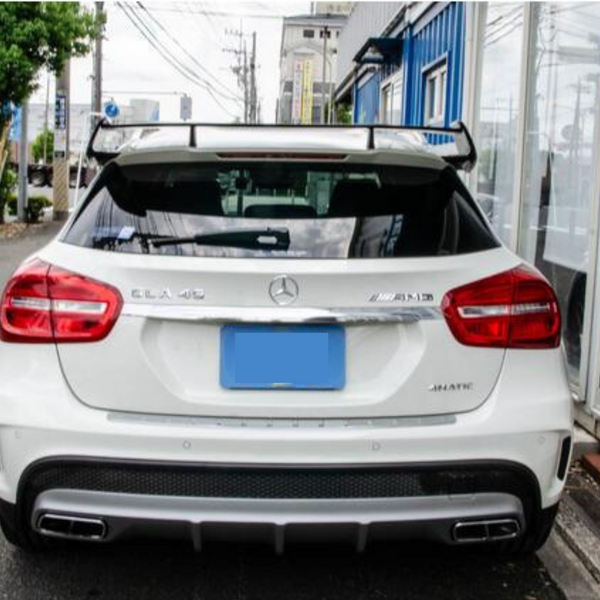 A white Mercedes-Benz GLA 45 AMG is parked on a street, with a distinctive spoiler. Nearby are trees, parked cars, and a blue building. Surrounding cables are visible in the sky.