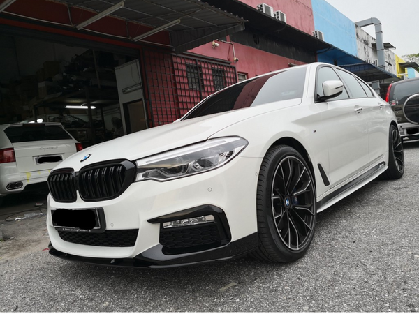 A white BMW sedan is parked on a gray asphalt surface outside a garage, surrounded by several other cars and colorful building walls.