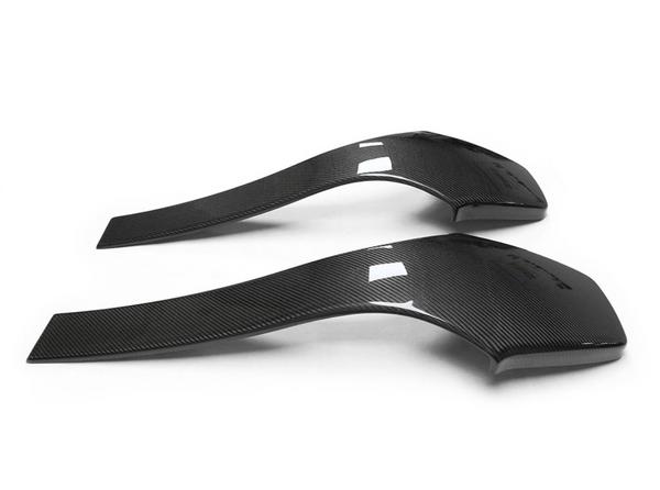 Two curved carbon fiber car spoilers rest side by side on a plain white background, exhibiting sleek, aerodynamic shapes with a glossy finish and subtle texture lines.
