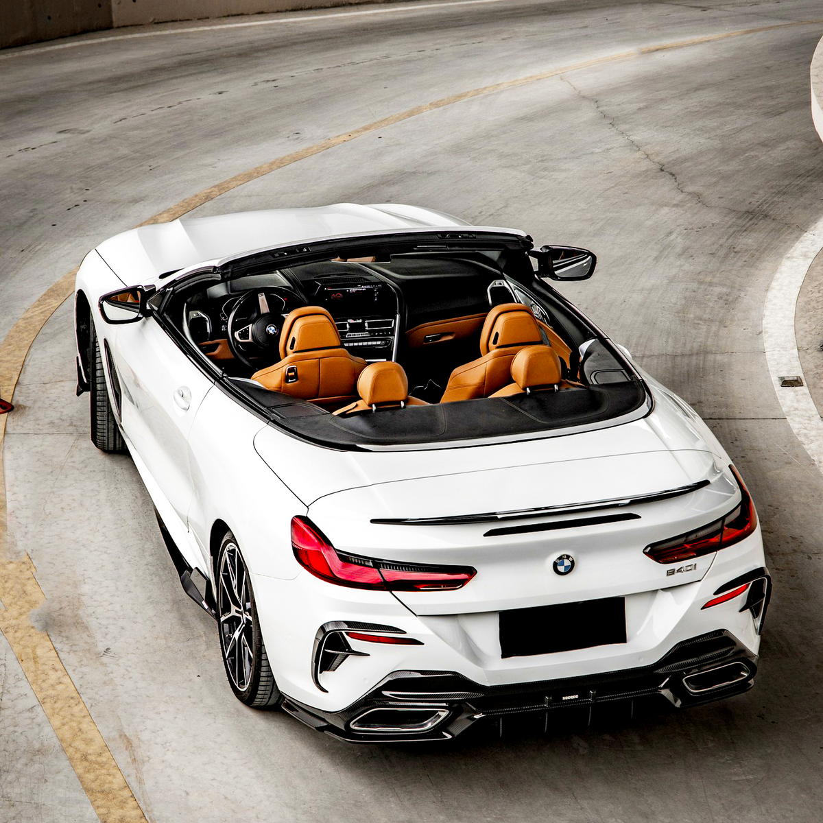 Convertible car is parked on a curving concrete road. The vehicle is white with a black soft top, showcasing tan leather seats. The surrounding area appears to be an industrial parking garage.