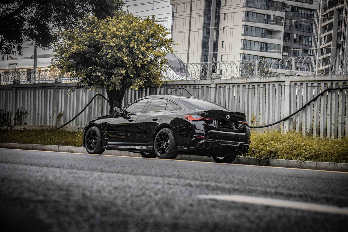A sleek black car is parked alongside a street, with a tree and a metal fence topped with barbed wire in the background, and high-rise buildings beyond.