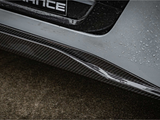 A carbon fiber car bumper with rain droplets glistening is shown against a wet pavement. Part of the word 