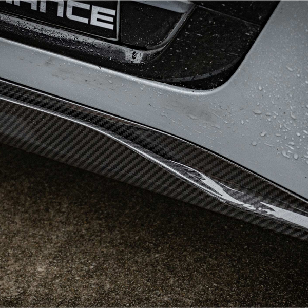 A carbon fiber car bumper with rain droplets glistening is shown against a wet pavement. Part of the word "ANCE" is visible on a nearby section.