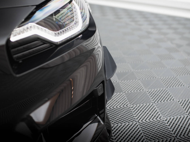A sleek black car's front headlight is illuminated, reflecting on its glossy surface. The car is parked on a patterned, gray-checkered floor, providing a modern backdrop.