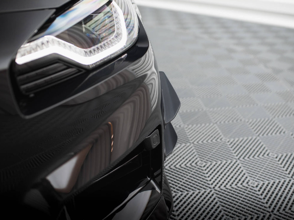 A sleek black car's front headlight is illuminated, reflecting on its glossy surface. The car is parked on a patterned, gray-checkered floor, providing a modern backdrop.