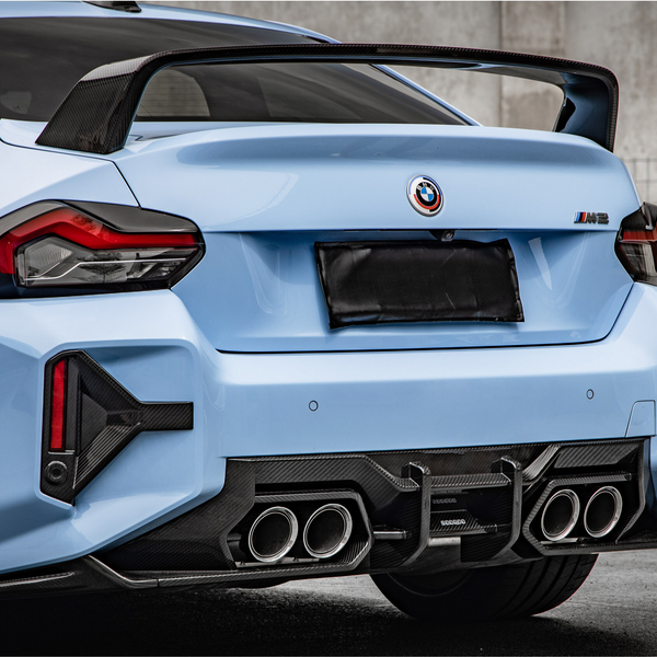 A blue car with a rear spoiler, four exhaust pipes, and a license plate obscured, parked on concrete with a metal fence and wall in the background.