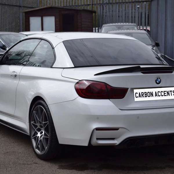 A white BMW M4 is parked on a lot with other cars, bordered by a fence. The license plate reads "CARBON ACCENTS." A small wooden shed is in the background.