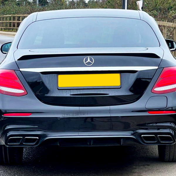 A black car is parked on a quiet road, aligned next to a wooden fence. The license plate is yellow, and a Mercedes-Benz logo is visible on the trunk.