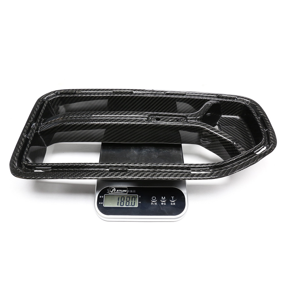 A carbon fiber car part rests on a digital scale, displaying "1180.0" grams. The scale has buttons labeled in another language, with a white background surrounding the setup.