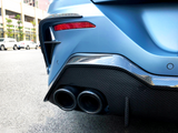 A blue car's dual exhaust pipes emit from a carbon fiber rear bumper on a city street, surrounded by parked white cars and a row of buildings.