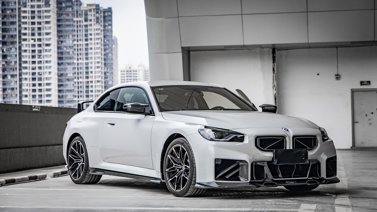A sleek white car is parked in a rooftop garage, surrounded by tall city buildings. It has modern, angular bodywork and black detailing, emphasizing its sporty design.