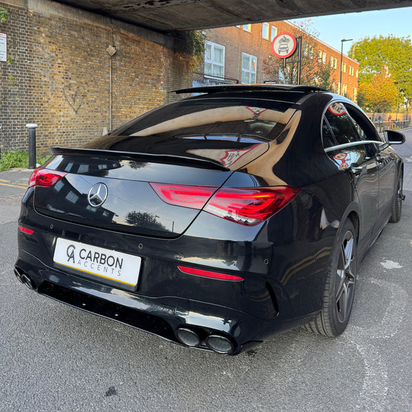 A black luxury car with a "Carbon Accents" plate is parked under a bridge. The surroundings include brick walls and buildings with trees under a clear sky.