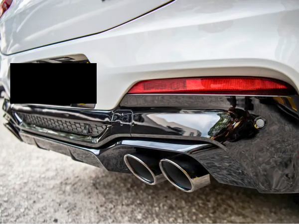 A white car's rear end is shown with dual exhaust pipes and a red taillight, parked on a gravel surface with another vehicle in the background.