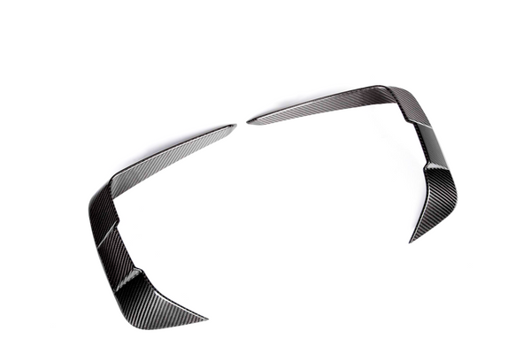 Two carbon fiber trim pieces, angled and sleek, lie on a plain white background, resembling modern automotive design elements, possibly for enhancing vehicle aerodynamics or styling.
