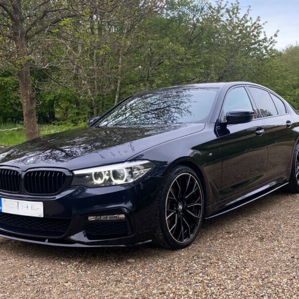 A black sedan with sleek design parked on a gravel path, surrounded by trees and greenery, under a clear sky. The car's headlights are on, reflecting the natural setting.