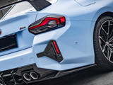 A light blue sports car displays a carbon fiber spoiler and quad exhausts, parked on an asphalt surface. The taillight design is angular and dramatic. Text on the bumper reads: 