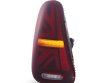 A sleek, triangular car taillight with a Union Jack design glows red and amber. It's positioned upright on a white reflective surface.