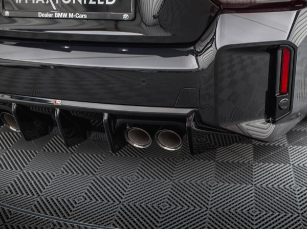 Car rear bumper with two exhaust pipes, parked on a checkered floor. A license plate above reads "MAXTONIZED Dealer BMW M-Cars."