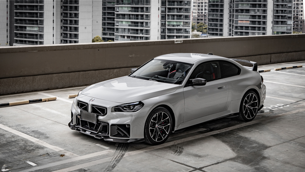 A sleek, silver sports car is parked on a rooftop, surrounded by tall urban buildings, under overcast skies.