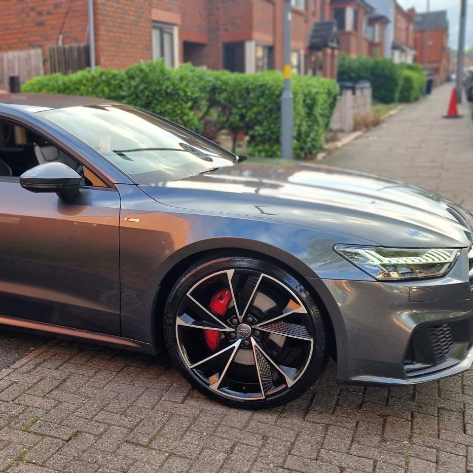 A sleek gray car is parked on a brick-paved street. Its front wheel features a distinctive alloy design. In the background, there are brick houses and greenery.