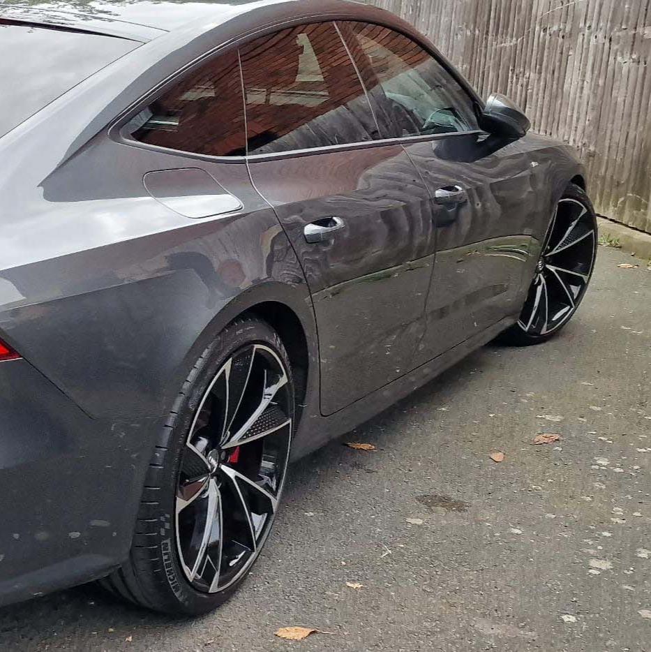 A grey car with black alloy wheels is parked on a paved surface beside a wooden fence, reflecting nearby buildings in its windows.
