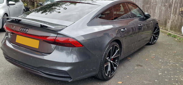 A gray Audi car is parked on a driveway beside a wooden fence. License plate is obscured, wheels are black with a unique design, and surroundings include another vehicle.