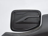 A black, rectangular fuel filler door sits on a white vehicle body. It features a sleek, geometric design with subtle ridges, surrounded by a slightly dusty surface.