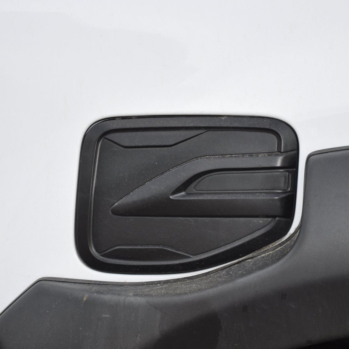 A black, rectangular fuel filler door sits on a white vehicle body. It features a sleek, geometric design with subtle ridges, surrounded by a slightly dusty surface.