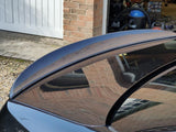 A black car with a carbon fiber spoiler is parked near a brick garage. The garage door is open, revealing various stored items, including a blue vacuum cleaner.