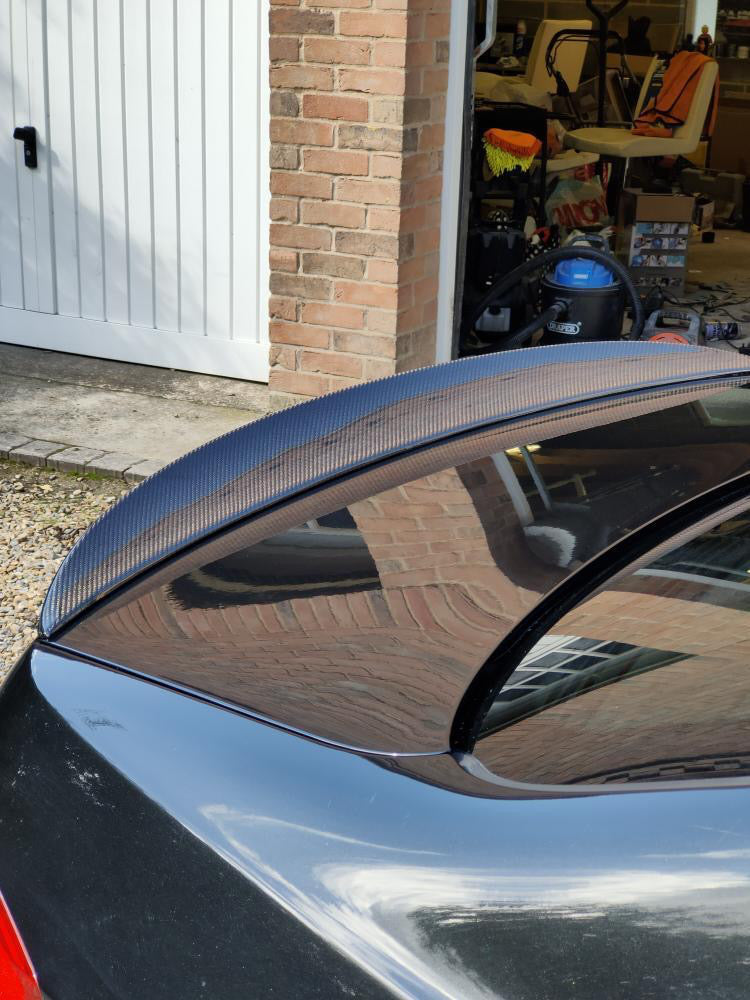 A black car with a carbon fiber spoiler is parked near a brick garage. The garage door is open, revealing various stored items, including a blue vacuum cleaner.