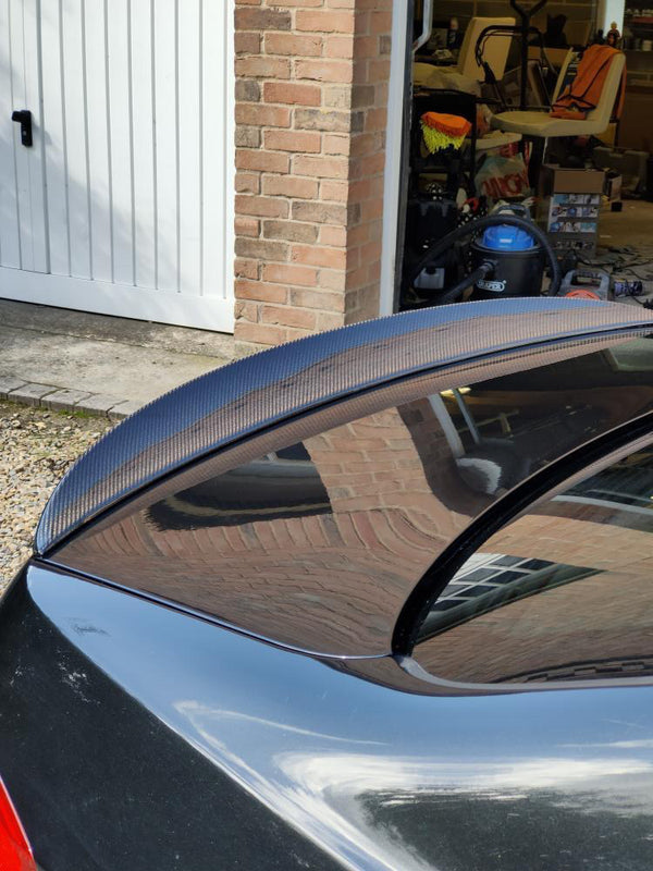 A black car with a carbon fiber spoiler is parked near a brick garage. The garage door is open, revealing various stored items, including a blue vacuum cleaner.