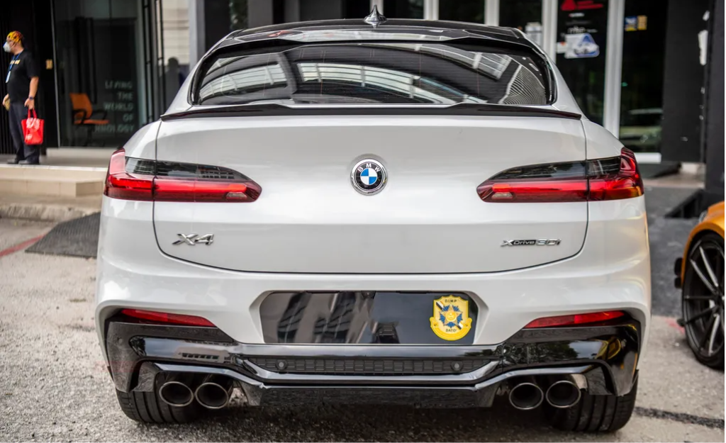 A white BMW X4 parked in front of a modern building with large glass doors. The car has dual exhausts and a badge labeled "xDrive 30i." There's a custom decorative plate.