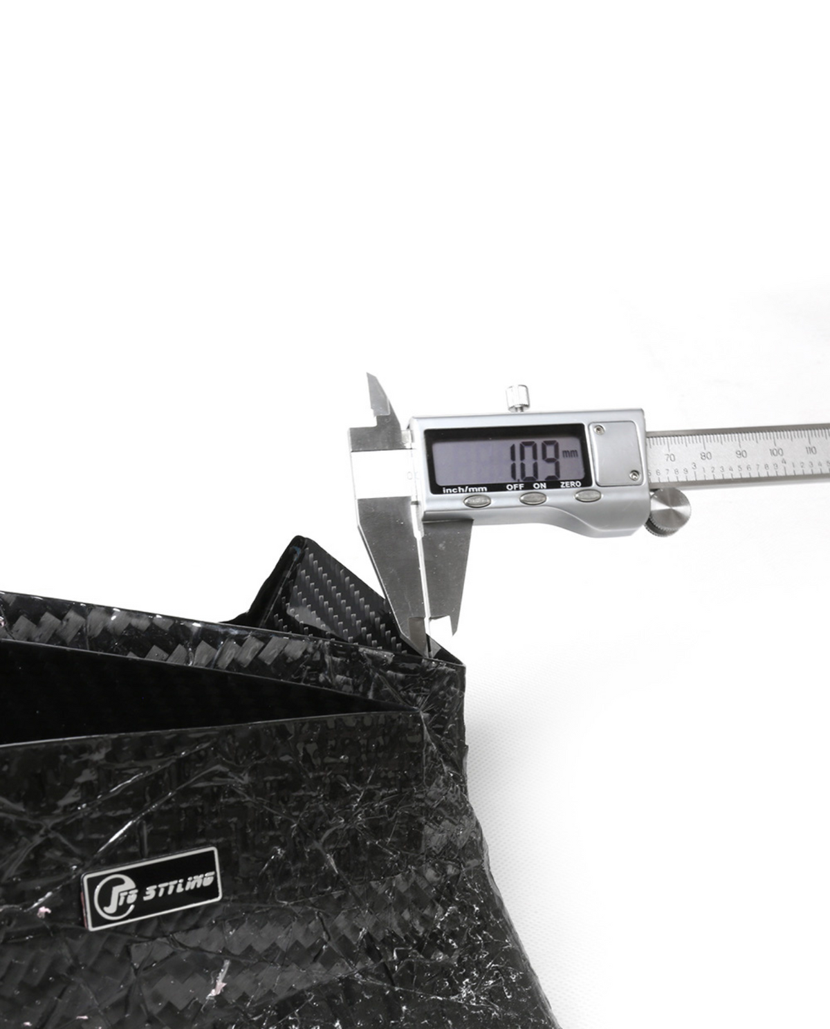 A digital caliper displays "10.9 mm" as it measures the thickness of a black carbon-fiber object. The setting is a clean, white background. Visible text: "Inch/mm OFF ON ZERO", "J's RACING".
