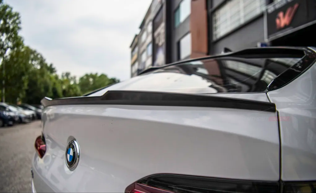 A white BMW car features a sleek rear spoiler, parked outdoors near a modern building with trees and more parked cars in the background.