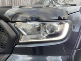 A car's headlight, reflecting nearby trees, is illuminated in daylight. It features a sleek design, situated next to a dark grille, with an 