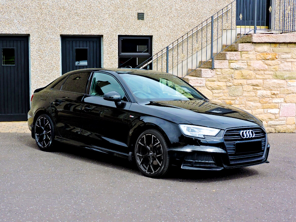 A black sedan car parked on an asphalt surface next to a building with stone walls and stairs, against a backdrop of closed doors.