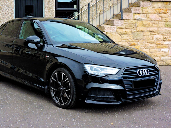 A sleek, black Audi car is parked on a paved surface, positioned beside a stone building with stairs and a door in the background.
