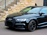A black Audi car is parked on a paved surface. Its sleek design is highlighted, positioned in front of a stone building with exterior stairs.