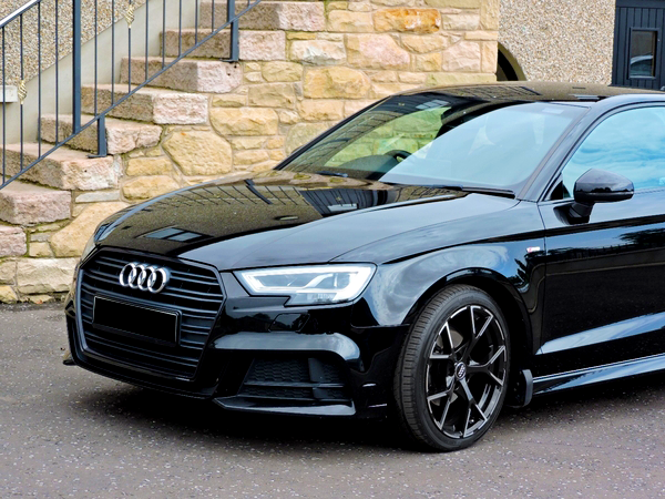 A black Audi car is parked on a paved surface. Its sleek design is highlighted, positioned in front of a stone building with exterior stairs.