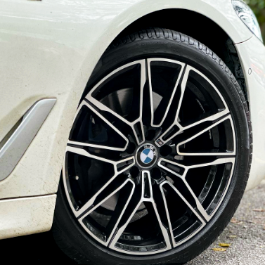 A car's wheel with a geometric alloy pattern and BMW logo is stationary on a paved surface, surrounded by part of the car's white fender and nearby ground.