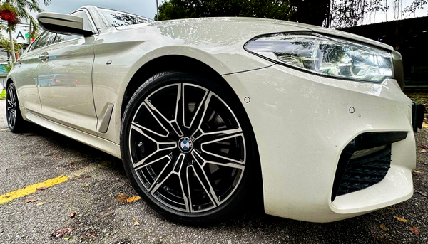 A sleek white BMW car, parked on a textured asphalt surface, features prominent black alloy wheels and illuminated headlights, surrounded by a few scattered leaves and greenery.
