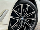 A BMW car tire with a detailed alloy rim design is stationary on a paved surface, partially surrounded by a white vehicle body and light foliage debris.