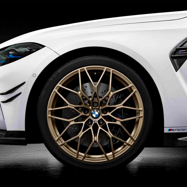 White sports car wheel features intricate gold alloy design with the BMW logo at the center. The car is parked indoors, highlighting details like "M Performance" on the body side.